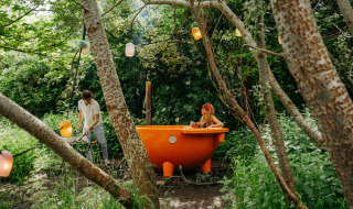Un hombre calienta una bañera naranja al aire libre mientras una mujer se relaja rodeada de bosque.