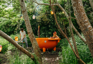 Un homme chauffe une baignoire orange en plein air où une femme se détend, entourés de nature luxuriante.