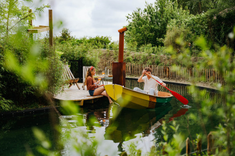 Man rows a colorful boat towards a woman relaxing on a wooden deck at Les Cabanes d'Ostende, Belgium.