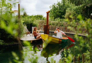Man rows a colorful boat towards a woman relaxing on a wooden deck at Les Cabanes d'Ostende, Belgium.