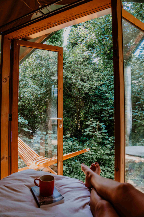 Persona relajada en la cama con café y libro, puerta abierta a la naturaleza en Les Cabanes d'Ostende, Bélgica.