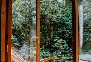 Persona relajada en la cama con café y libro, puerta abierta a la naturaleza en Les Cabanes d'Ostende, Bélgica.