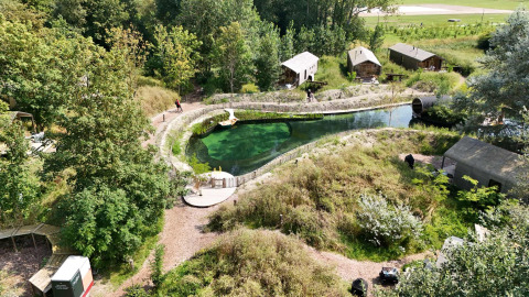 Luftfoto af Les Cabanes d'Ostende feriepark med grønne træer, små hytter og naturlig swimmingpool.