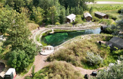 Luftfoto af Les Cabanes d'Ostende feriepark med grønne træer, små hytter og naturlig swimmingpool.