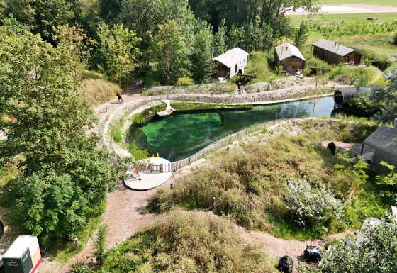 Luftfoto af Les Cabanes d'Ostende feriepark med grønne træer, små hytter og naturlig swimmingpool.