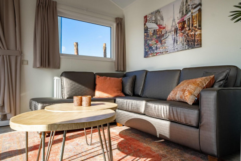 Wohnzimmer mit dunklem Sofa, runden Tischen, Wandbild und Sonnenlicht in der Velthorst-Lodge, Zilverstrand.