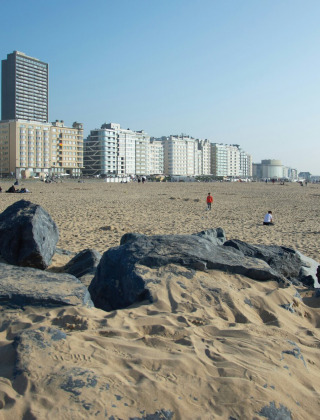 Playa de arena con rocas en Oostende, Bélgica, edificios residenciales altos al fondo en un día soleado.