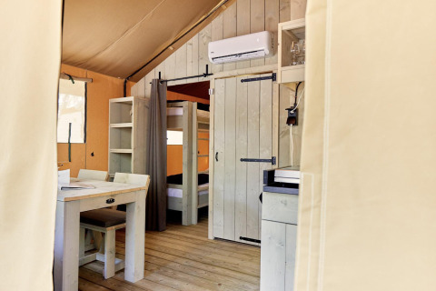 Vista interna di una tenda glamping con letti a castello, tavolo da pranzo, scaffale e aria condizionata a parete.