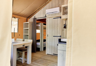 Vista interna di una tenda glamping con letti a castello, tavolo da pranzo, scaffale e aria condizionata a parete.