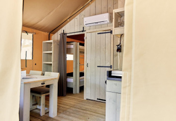 Vista interna di una tenda glamping con letti a castello, tavolo da pranzo, scaffale e aria condizionata a parete.