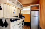 Moderne Glampingküche mit Holzwänden, Herdplatte, offenem Regal und silbernem Kühlschrank.