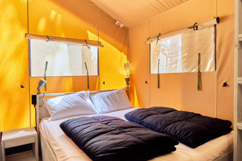 Alojamiento glamping con cama doble, almohadas blancas, edredones oscuros y luz natural entrando por ventanas.