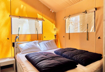Hébergement glamping avec lit double, oreillers blancs, couettes noires et lumière naturelle par les fenêtres.