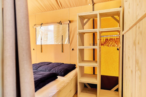 Interno di una tenda glamping con letto, scaffale in legno e appendiabiti in un ambiente luminoso.