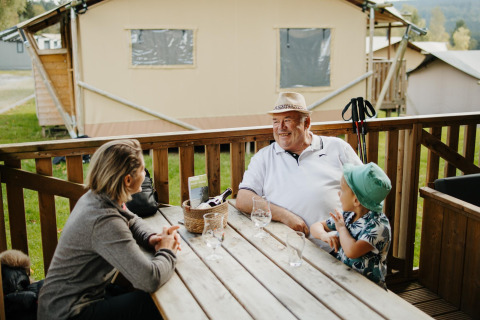 Tres personas conversan en la terraza de madera en la cabaña Sunshine de Petite Suisse, Bélgica.