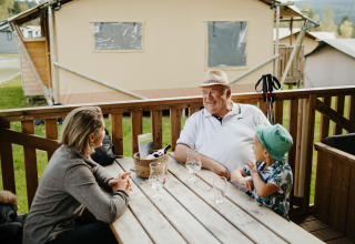 Tres personas conversan en la terraza de madera en la cabaña Sunshine de Petite Suisse, Bélgica.