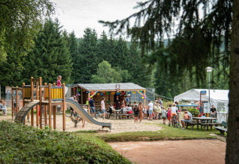 Des enfants jouent sur une aire de jeux près du lodge Sunshine à Petite Suisse, Belgique, avec des familles autour.