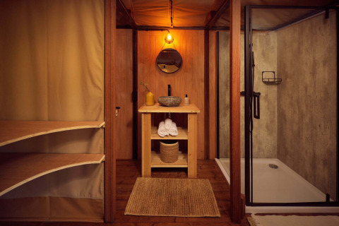 Baño en una tienda de glamping safari con elementos de madera, ducha moderna y decoración rústica natural.