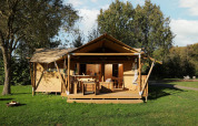 Safari tent with a covered wooden porch on grassy lawn, surrounded by trees under blue sky and sun.