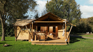 Tienda safari con porche cubierto de madera, en césped, rodeada de árboles y cielo azul soleado.
