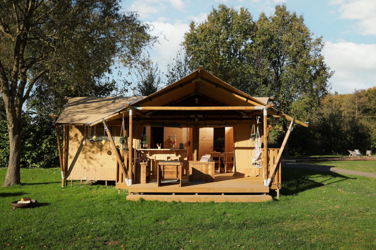 Safari tent with a covered wooden porch on grassy lawn, surrounded by trees under blue sky and sun.
