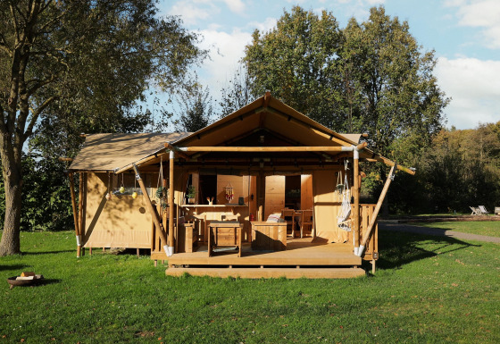 Tenda safari con veranda in legno coperta su prato verde, circondata da alberi e cielo azzurro.
