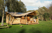 Tienda safari con porche de madera, silla colgante y área de descanso, rodeada de césped y árboles verdes.