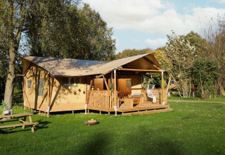 Safarizelt mit Holzveranda und Sitzgelegenheiten, umgeben von Bäumen und einer grünen Wiese im Sonnenlicht.