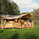 Safari tent with a wooden porch, swing chair, and seating area, set in a green grassy field with trees.