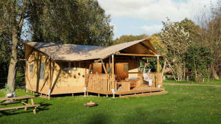 Tienda safari con porche de madera, silla colgante y área de descanso, rodeada de césped y árboles verdes.