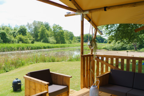 Udsigt fra en glamping-teltterrasse ved en sø, med træmøbler og grøn natur i Resort de Parel, Holland.