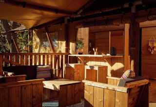 Terrasse de tente safari avec mobilier en bois, coussins colorés et ambiance chaleureuse en pleine nature.