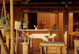 Intérieur de tente safari avec mobilier en bois, comptoir de bar, plantes suspendues et lumière douce.