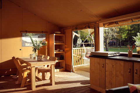 Interior photo of a safari tent kitchen and dining area with wooden furniture, terrace, and nature view.