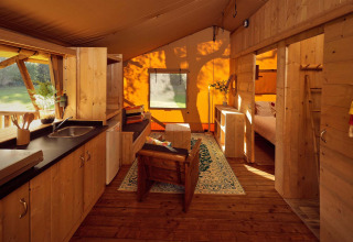 Intérieur d'une tente safari de luxe avec mobilier en bois, kitchenette et chambre éclairée par le soleil.