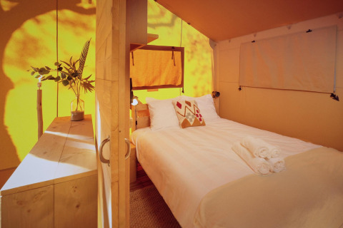 Interior de una tienda safari glamping con cama doble, muebles de madera y luz natural cálida entrando.