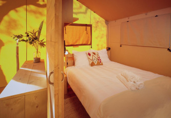 Interior of a glamping safari tent with a cozy bed, wooden furniture, white linens and warm natural sunlight.