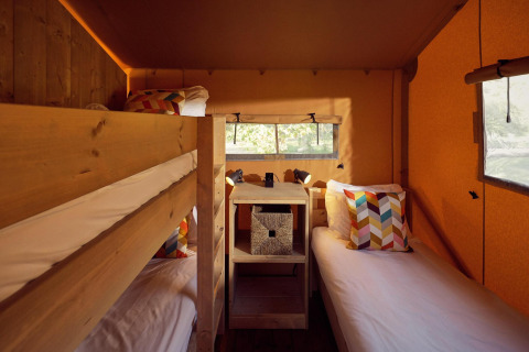Intérieur d'une tente safari avec lits superposés, lit simple, coussins colorés et murs en bois clair.