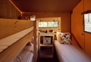 Intérieur d'une tente safari avec lits superposés, lit simple, coussins colorés et murs en bois clair.