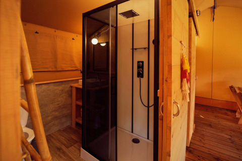 Vue intérieure d'une tente safari avec une douche moderne, un sol en bois et un éclairage chaleureux.