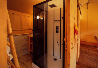 Vista interna di una tenda safari con cabina doccia moderna, pavimento in legno e luce calda.