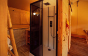 Vue intérieure d'une tente safari avec une douche moderne, un sol en bois et un éclairage chaleureux.