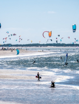 Playa llena de kitesurfistas y cometas junto a un parque vacacional ideal para glamping moderno.