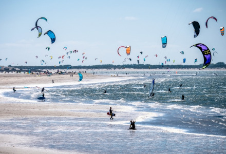 Playa llena de kitesurfistas y cometas junto a un parque vacacional ideal para glamping moderno.