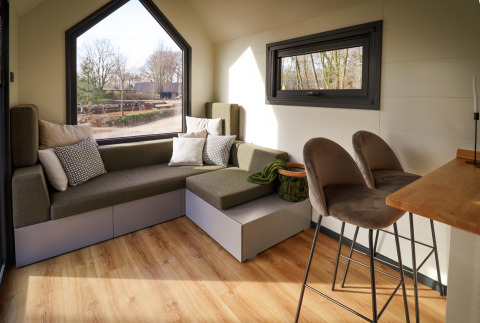 Intérieur d'une tiny house au Holiday Park Sallandshoeve avec canapé, tabourets de bar et grandes fenêtres.