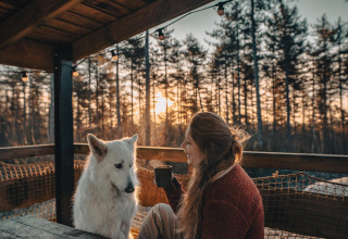 Vrouw geniet van een warme drank met haar witte hond op het terras bij Treetrunk lodge, Cosy Cabins, Limburg, België.