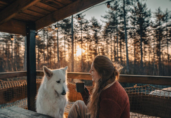 Kvinde nyder en varm drik med sin hvide hund på terrassen ved Treetrunk lodge, Cosy Cabins, Limburg, Belgien.