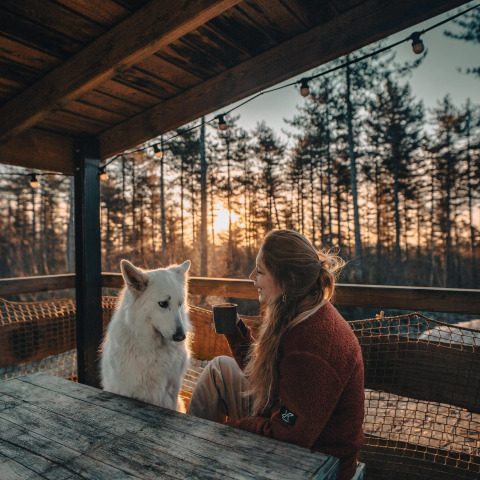 Kvinde nyder en varm drik med sin hvide hund på terrassen ved Treetrunk lodge, Cosy Cabins, Limburg, Belgien.