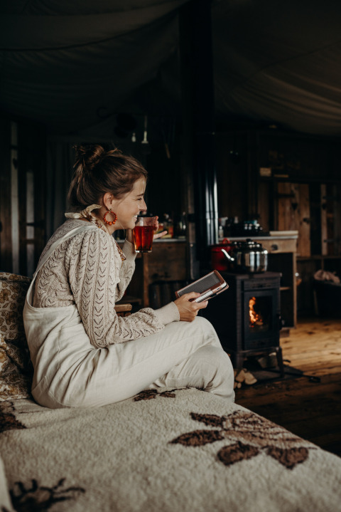 Vrouw geniet van een warme drank en leest bij de houtkachel op Feather Down TaarTenTuin, Zuid-Holland.