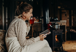 Femme dégustant une boisson chaude et lisant un livre près du poêle à Feather Down TaarTenTuin, Hollande-Méridionale.